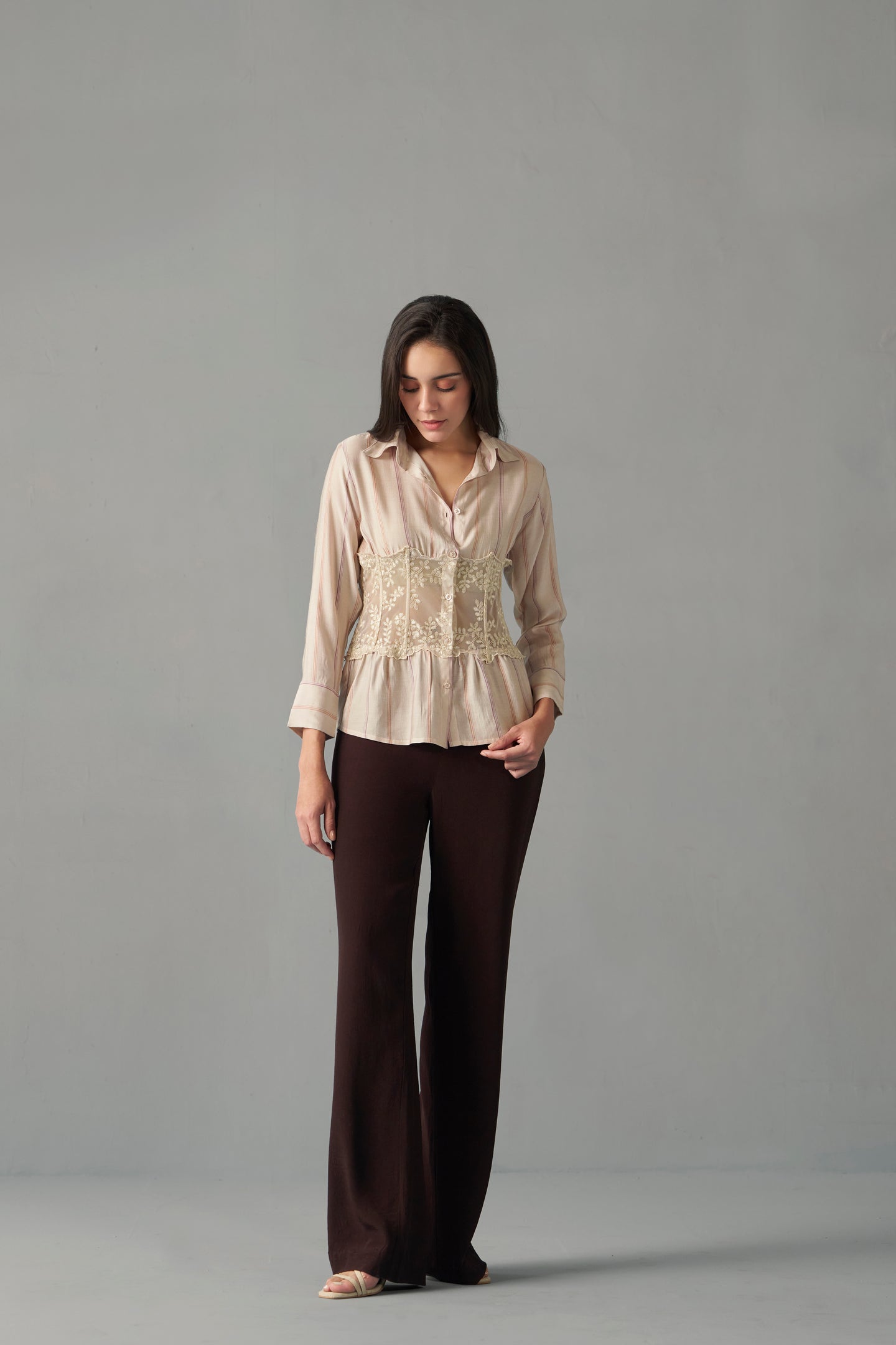 Model wearing embroidered corset detail stripe print shirt , Woman wearing a beige blouse with lace details and brown pants on a gray background