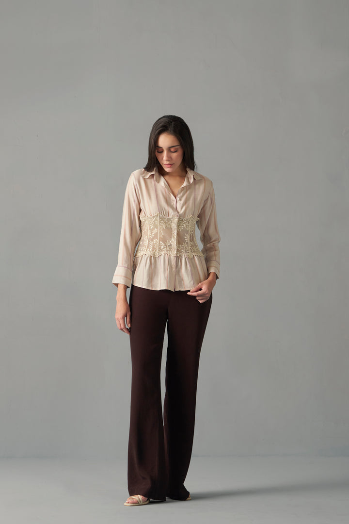 Model wearing embroidered corset detail stripe print shirt , Woman wearing a beige blouse with lace details and brown pants on a gray background