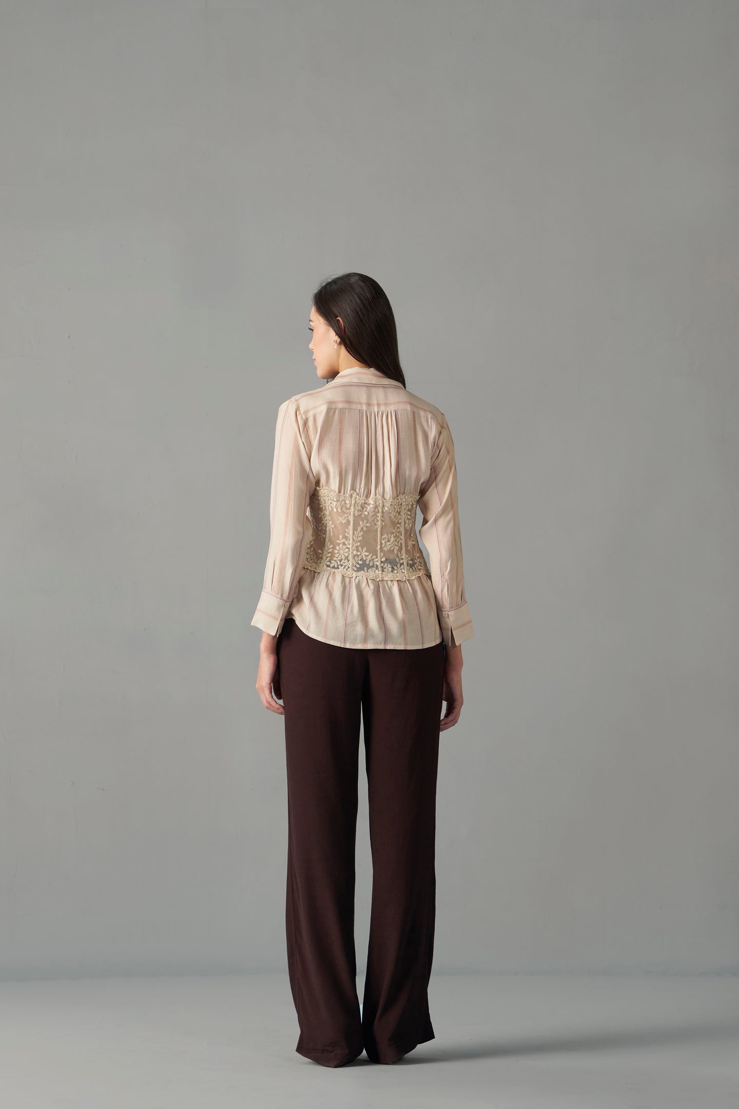 Embroidered corset detail shirt back view with tailored fit, Woman wearing a beige blouse with lace details and brown pants on a gray background